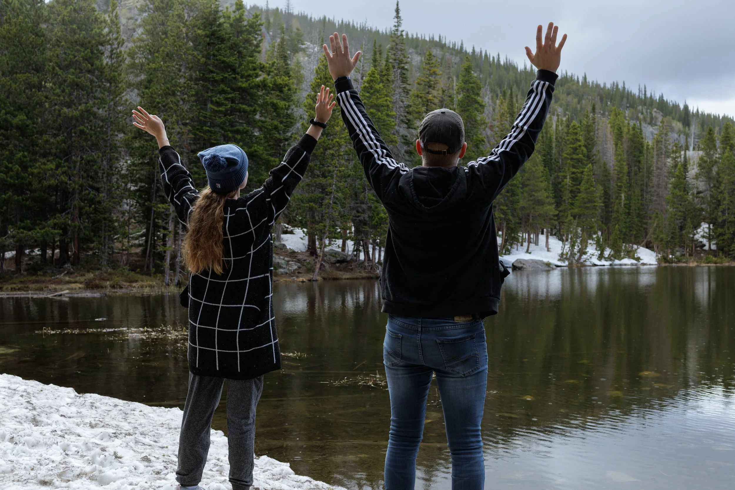 Two people, one girl and one boy, standing by a lake in a forested mountain area, raising their arms in celebration or greeting.