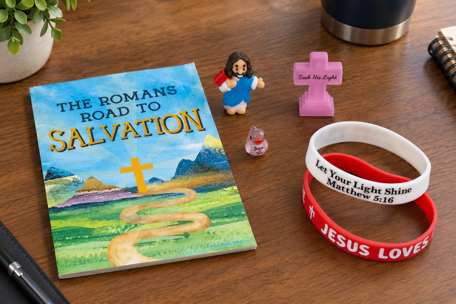 Christian religious items on a wooden table, including a booklet titled 'The Romans Road to Salvation,' a figurine of Jesus carrying a cross, a small piggy bank with 'Jesus You' written on it, a pink cross with 'Seek His Light' inscribed, and two bracelets with religious messages.