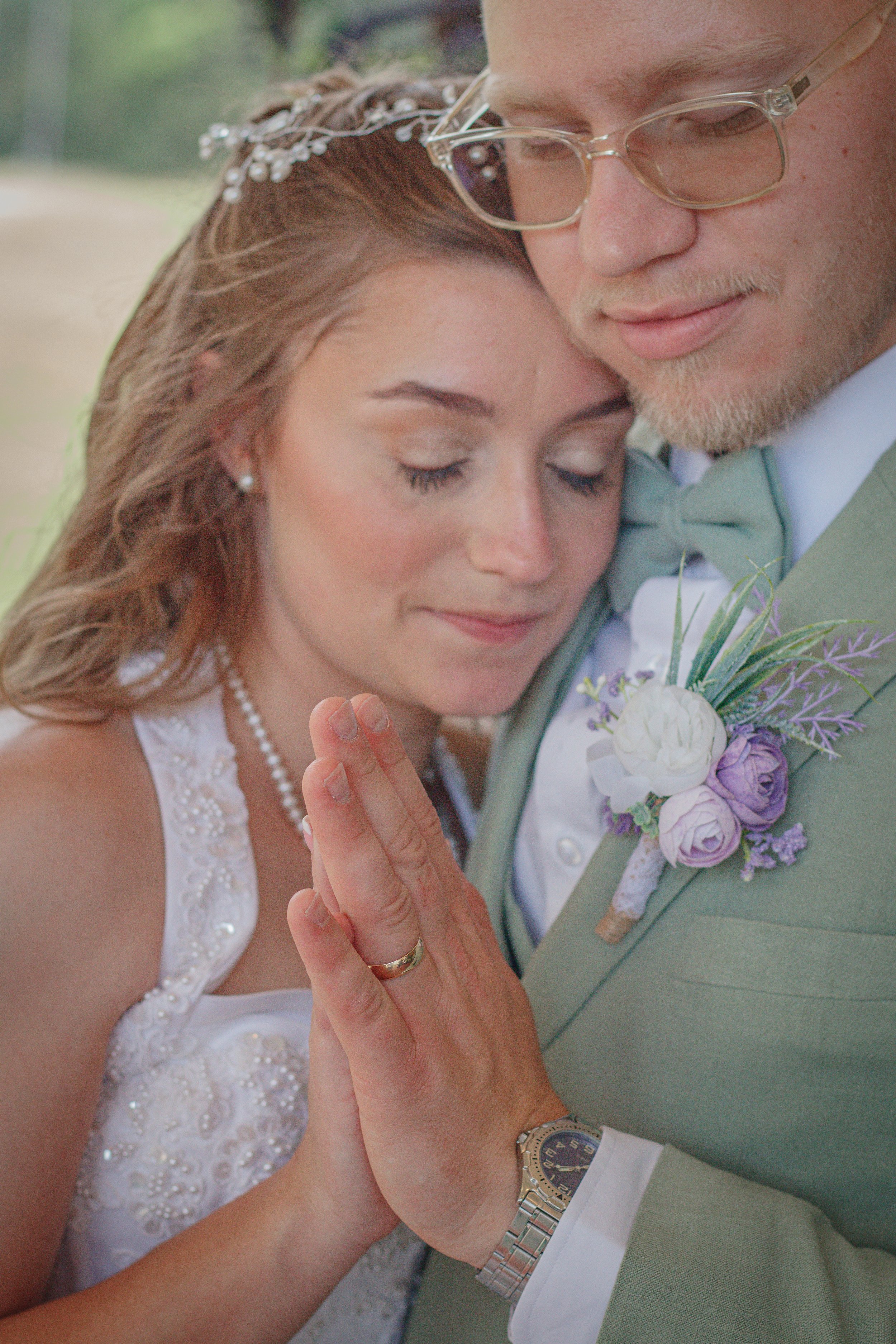 A bride and groom share a tender moment with their foreheads touching and eyes closed, during their wedding ceremony. The bride is wearing a pearl necklace and a white lace dress, while the groom is in a light green suit with a floral boutonniere, glasses, and a silver wristwatch.
