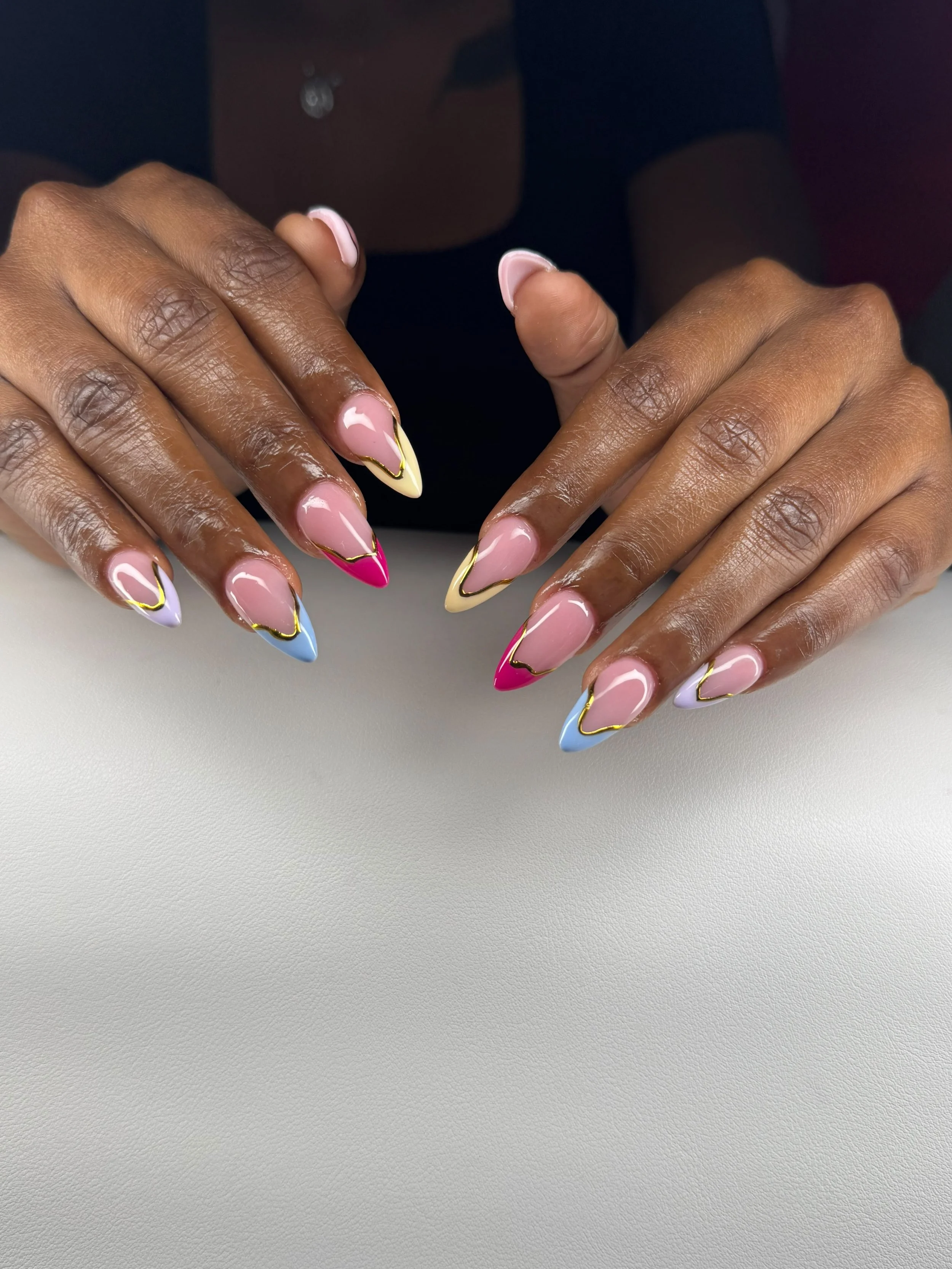 Close-up of a person's hands with pastel-colored almond-shaped nails featuring a yellow and gold outline design, resting on a white surface.
