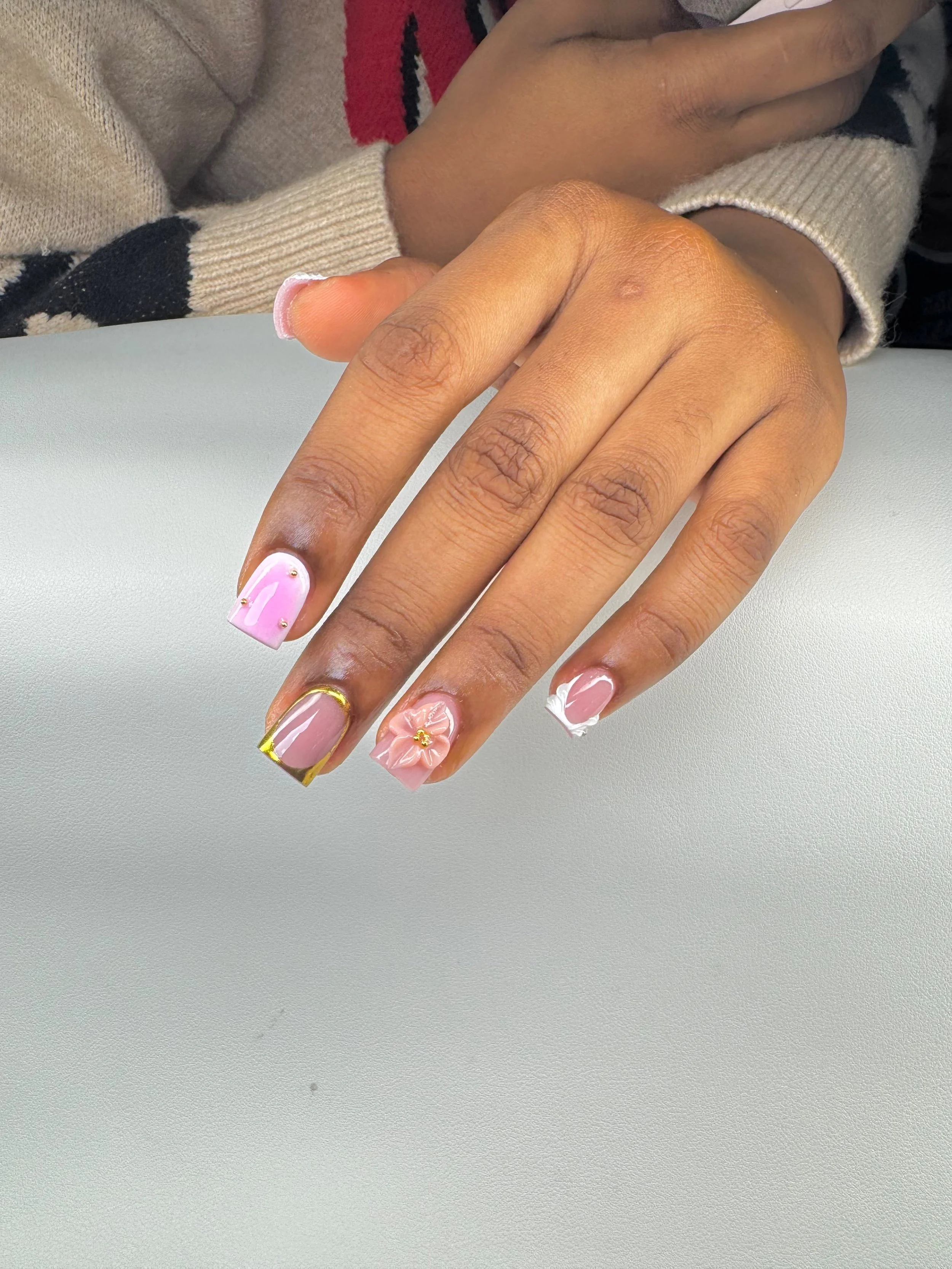 Close-up of a woman's hand with pressed-on nails featuring different designs, including pink with gold accents, pink with a flower, and pink with white tips, resting on a light gray surface. A person is in the background with crossed arms wearing a beige and black sweater.