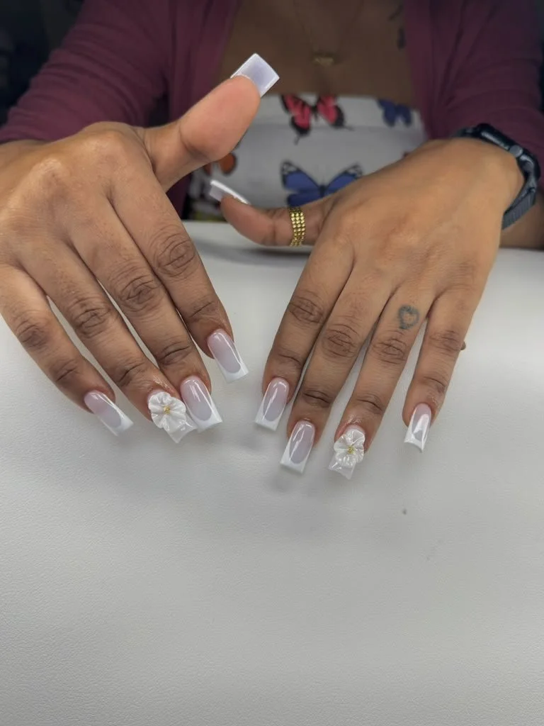 Close-up of a woman showing her freshly manicured nails with white French tips and 3D white flower nail art, on a white surface.