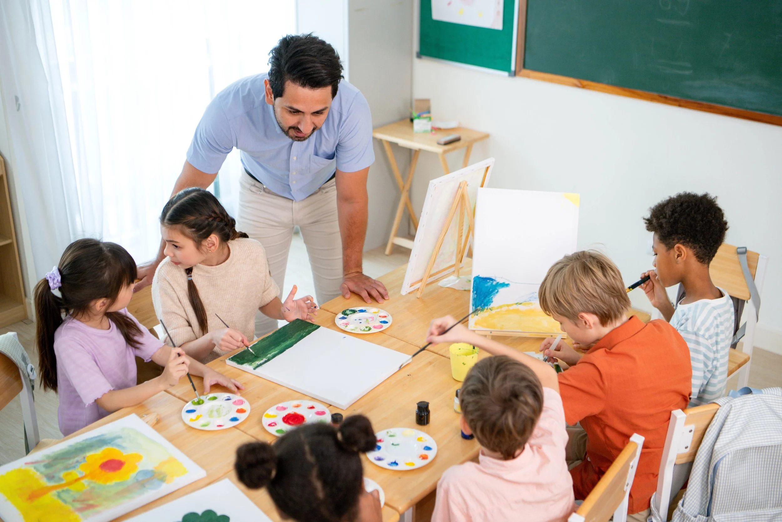 A teacher guiding children in a classroom during a painting activity, with children painting on paper and supplies on the table.