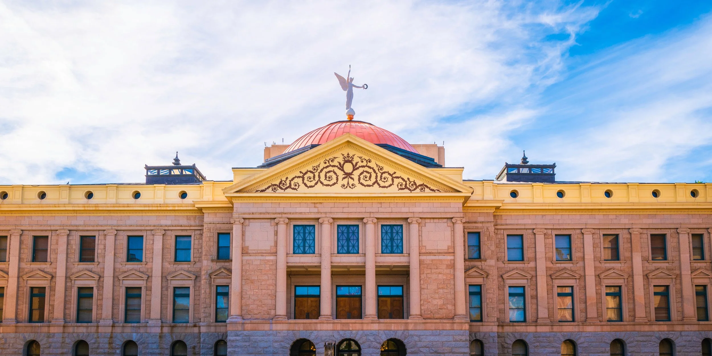 The facade of a historic building with classical architecture, featuring columns, decorative details, and a pediment with an ornate design. A dome with a statue on top is visible against a partly cloudy sky.