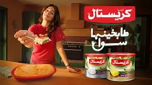 A woman in a red shirt holding a plate of food in a kitchen with a box of Crestistal yogurt containers in the foreground.