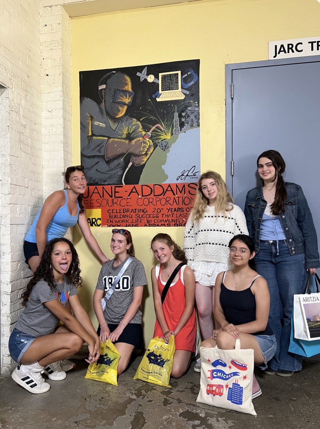 Group of seven young women posing indoors in front of a colorful mural celebrating Jane Addams Resource Corporation, with some holding yellow and one holding a tote bag with Chicago-themed prints.