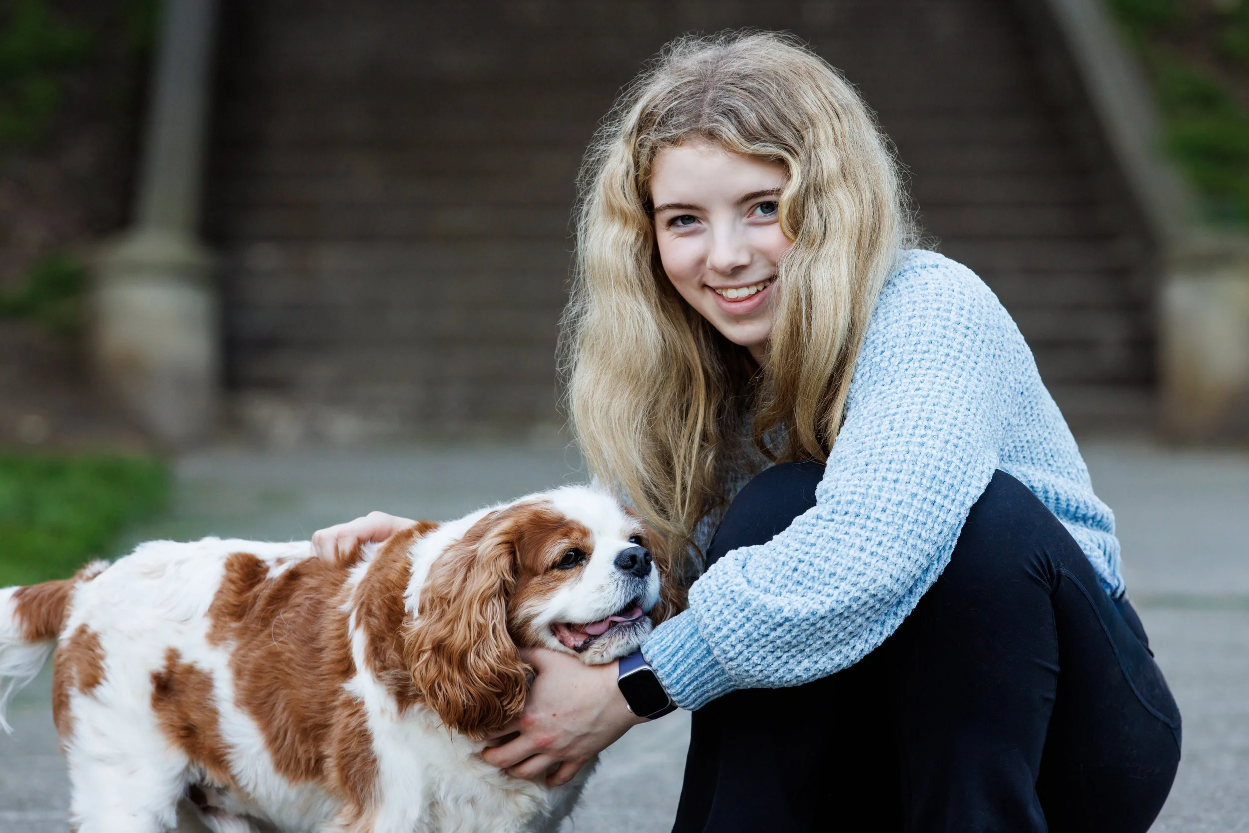 A girl with long blonde hair, smiling, hugging a Cavalier King Charles Spaniel dog outdoors near stairs.