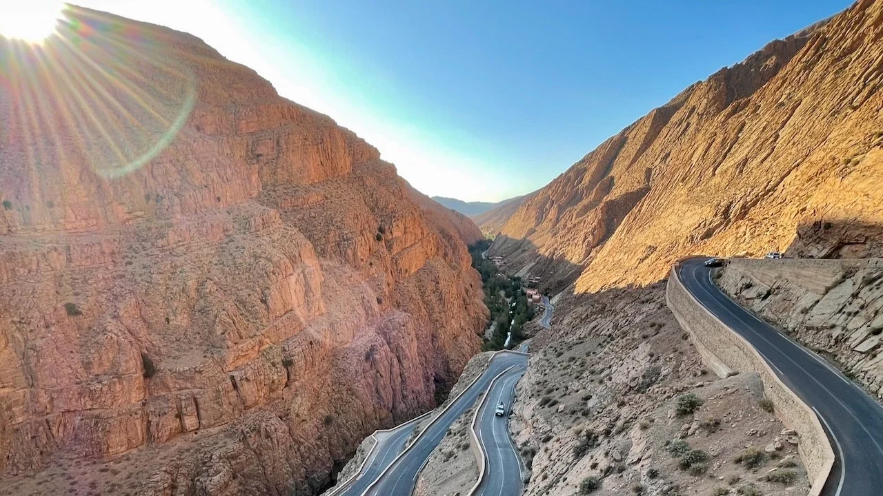 A winding mountain road through a steep, sunlit red-rock canyon.
