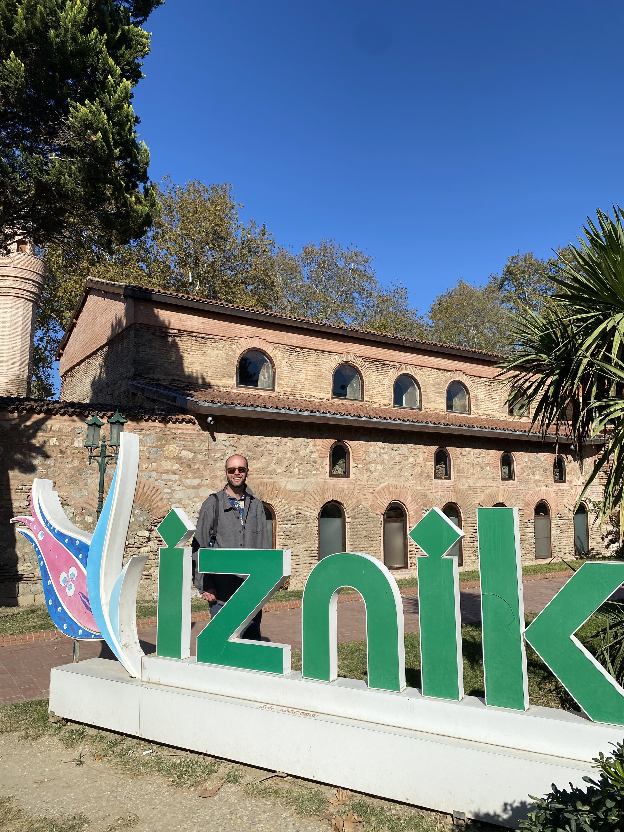 Jacob standing beside an İznik sign in front of a historic stone building under a bright blue sky.