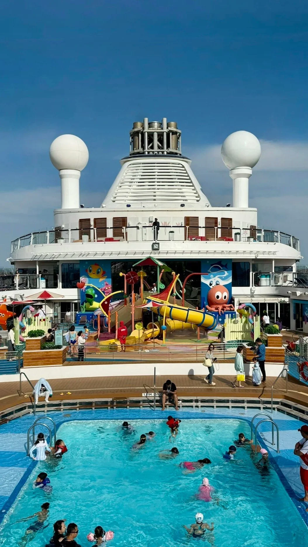 A large cruise ship pool deck with swimmers, waterslides, and outdoor activities.