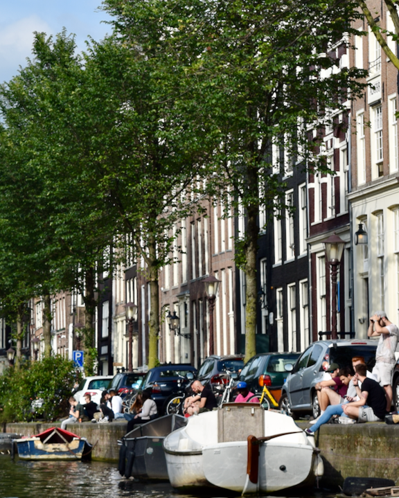 Amsterdam canal lined with narrow houses, trees, houseboats, and people sitting along the waterfront.