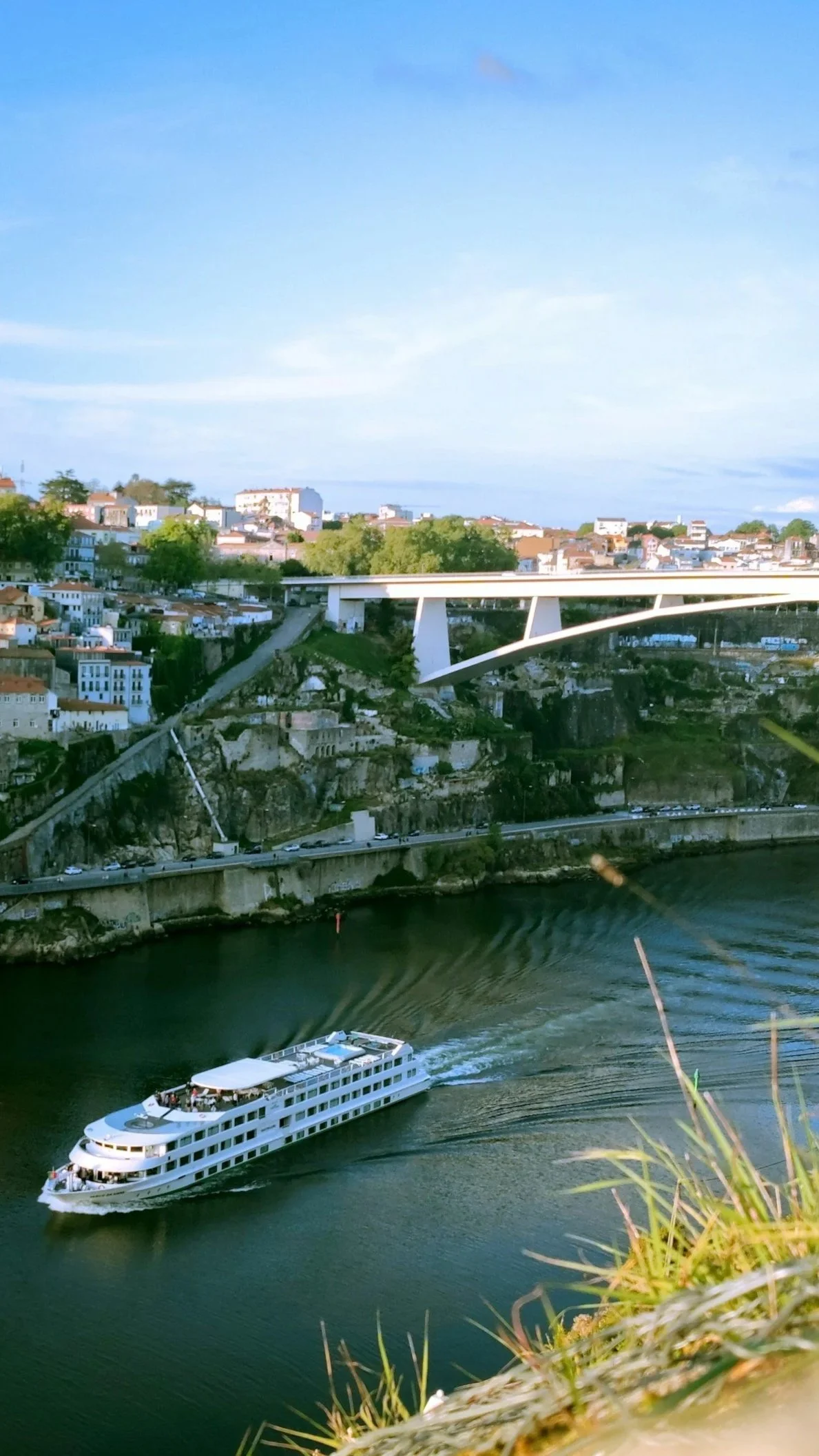 A river cruise ship sailing past a European city beneath a high bridge.