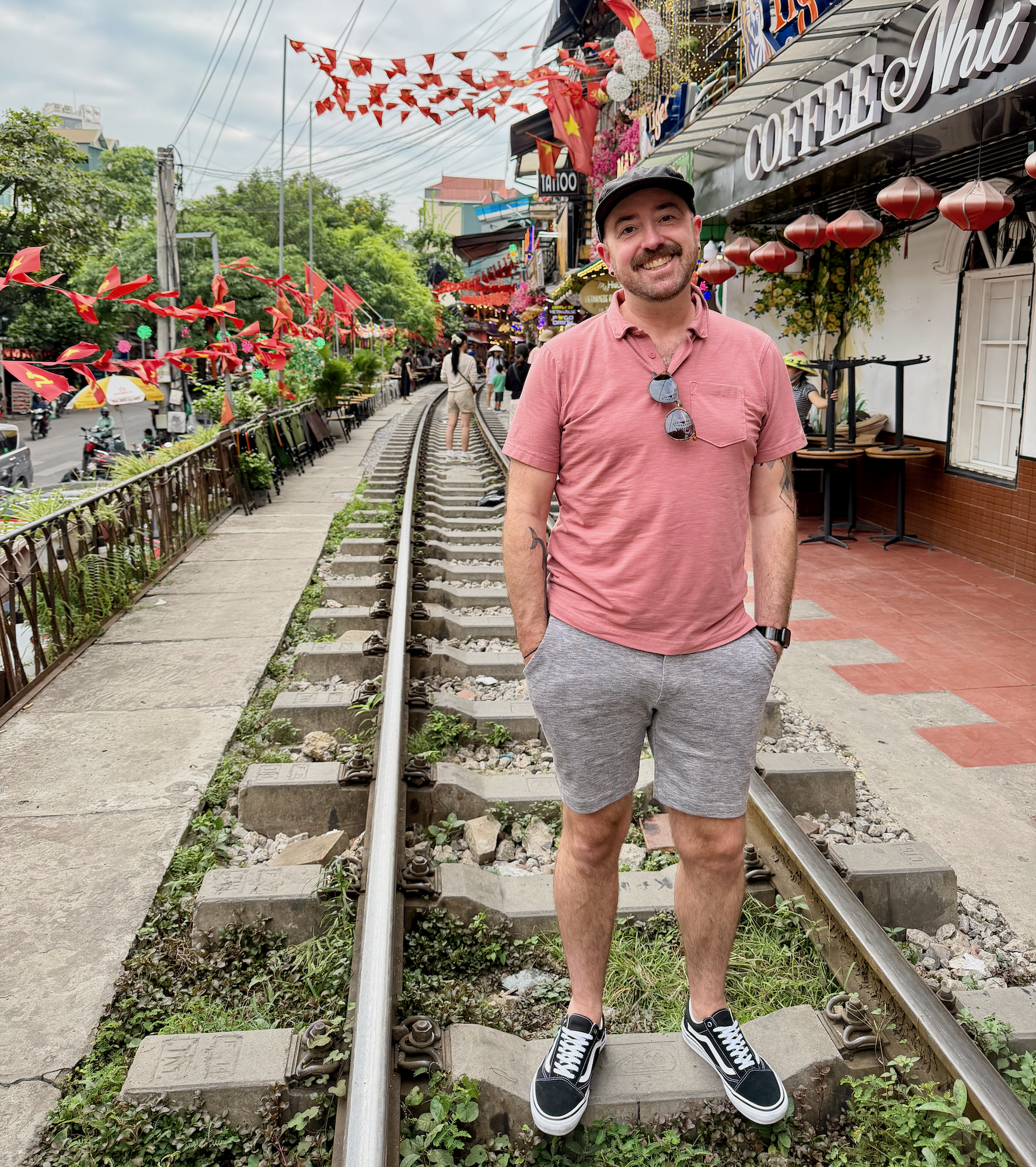 A traveler standing on railway tracks in Hanoi along a narrow street lined with cafés, lanterns, and greenery.