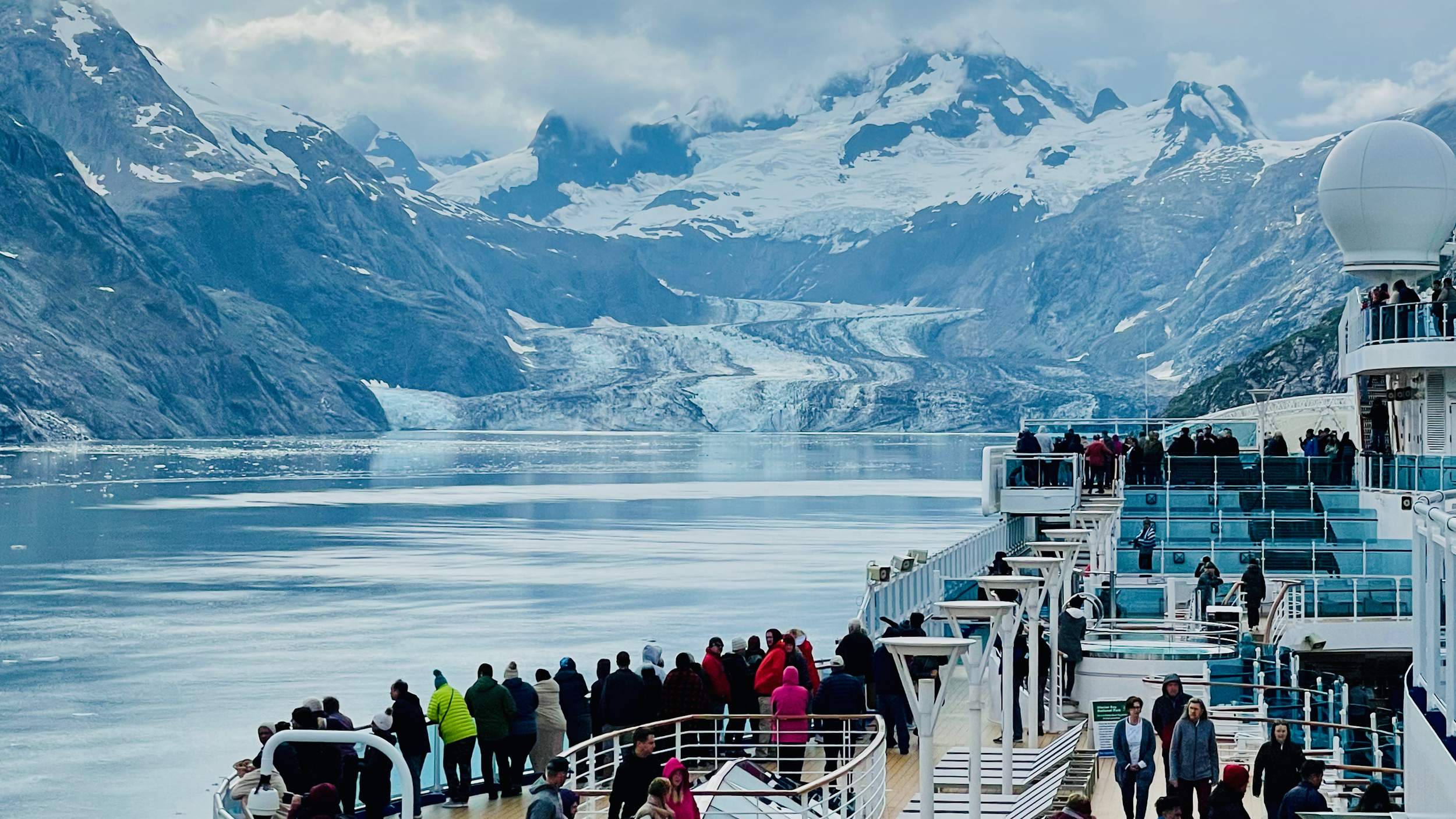 Cruise passengers on deck facing a glacier surrounded by snow-covered mountains.