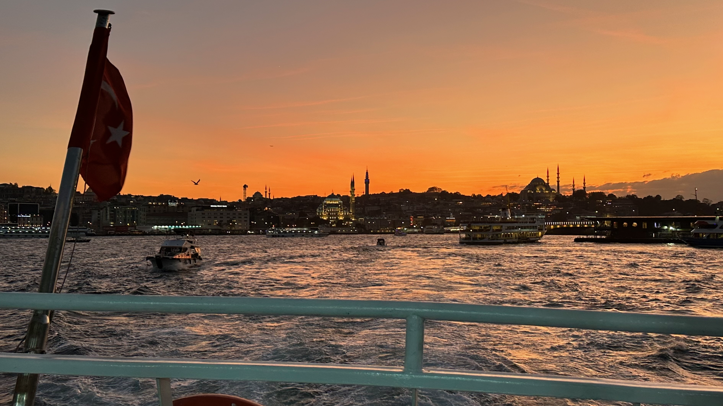 Sunset over Istanbul from the water, with a Turkish flag in the foreground and mosque minarets on the skyline.