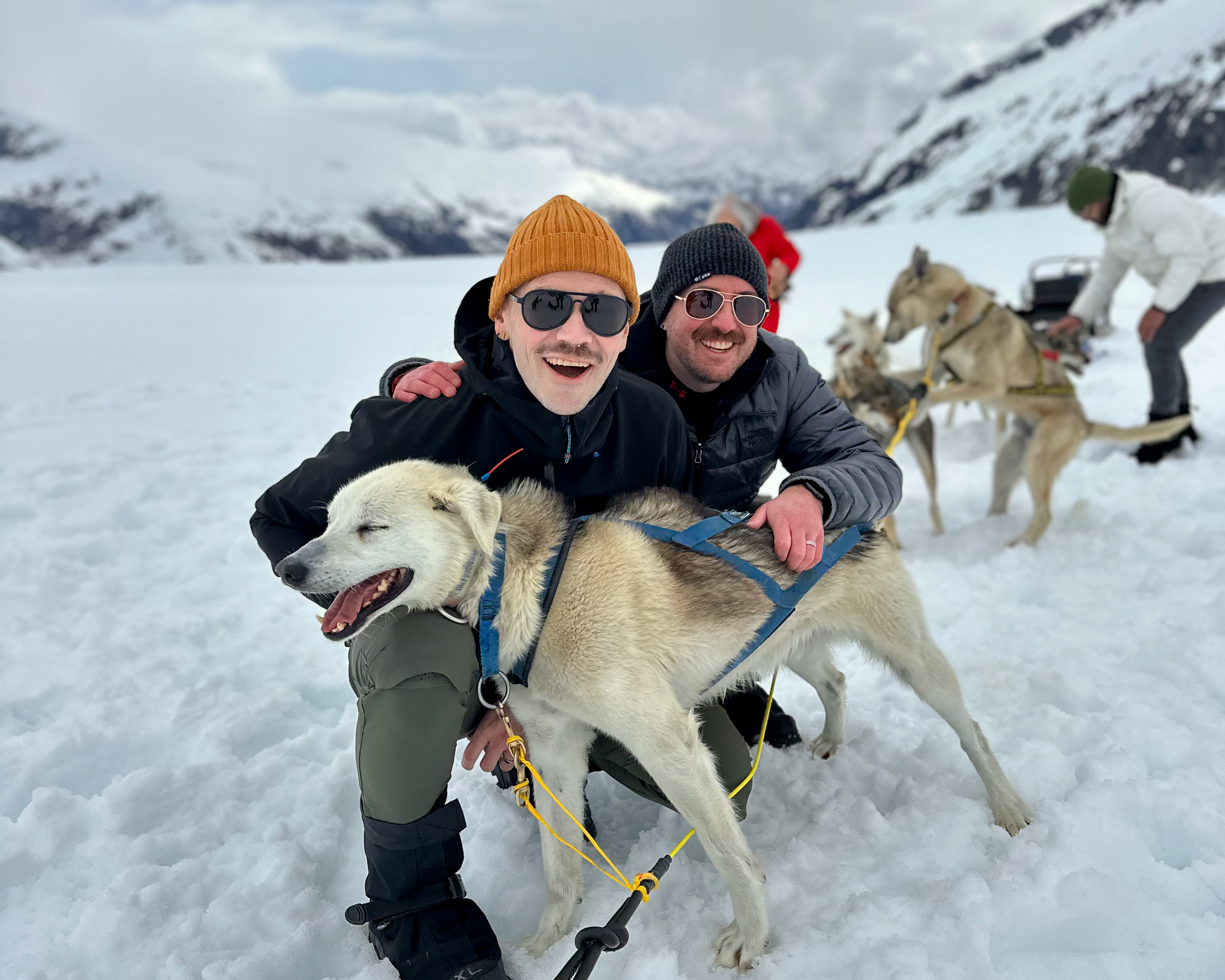 Two travelers smiling beside a sled dog in a snowy mountain landscape.