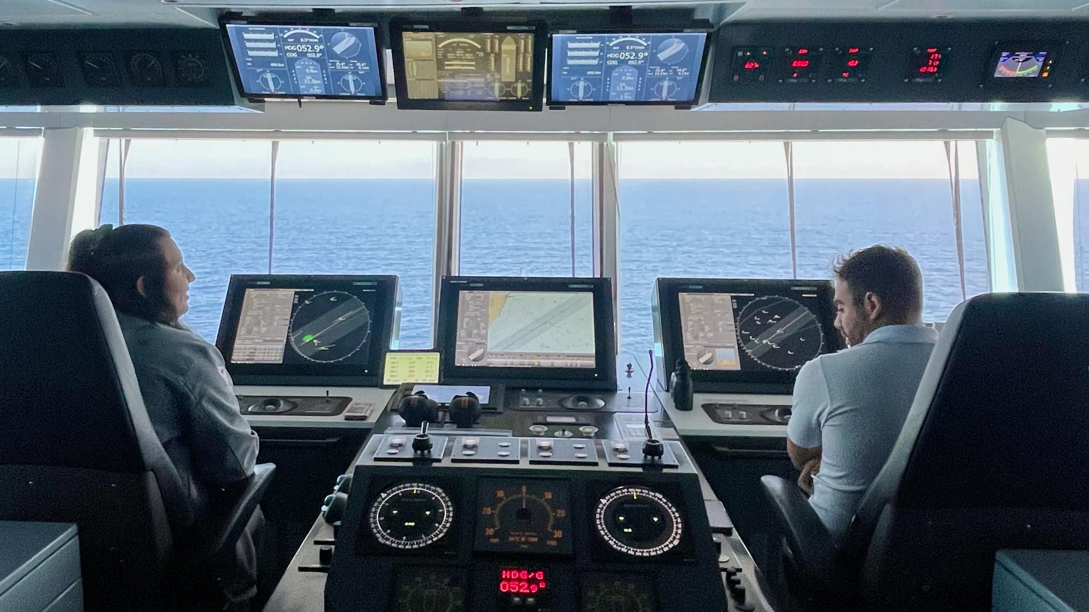 Two officers at the navigation consoles on the bridge of a cruise ship, with the ocean visible ahead.