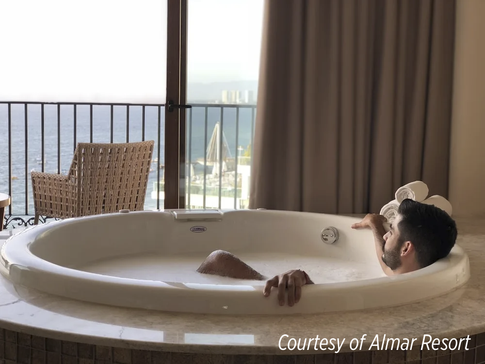 A hotel guest relaxing in a large in-room soaking tub with an ocean view.