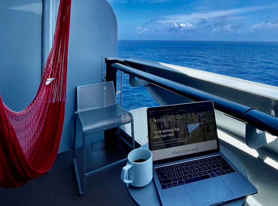 A cruise-ship balcony set up for a quiet sea day, with a hammock, laptop, coffee, and open ocean beyond.