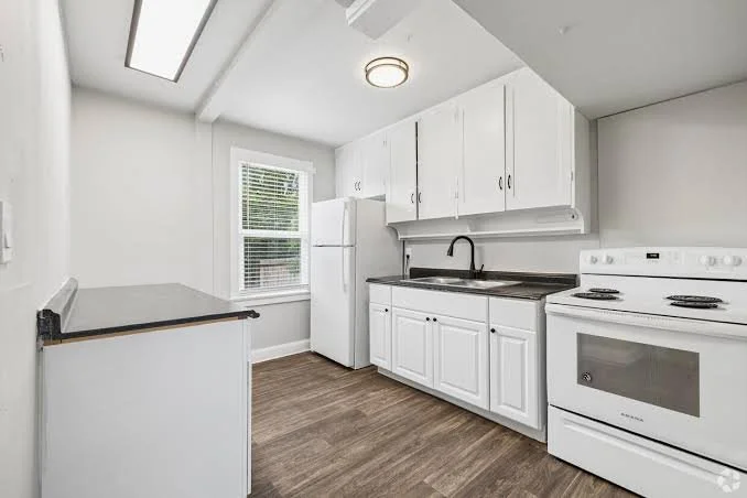 Kitchen with white cabinets, refrigerator, stove, and window, with wood flooring and ceiling light.
