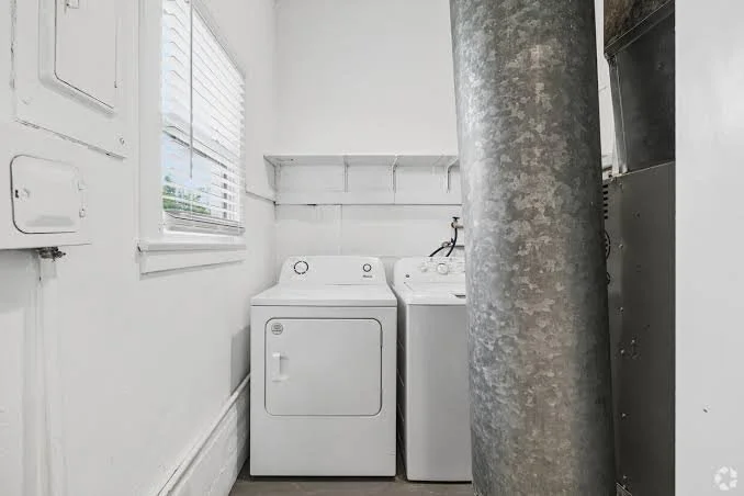 Small laundry room with white washing machine and dryer, white walls, window with blinds, and a large metal pipe.