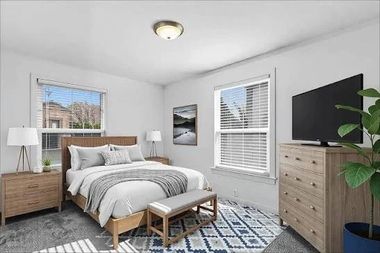 A bedroom with a large bed, two nightstands with lamps, a dresser with a TV, and a window with blinds.