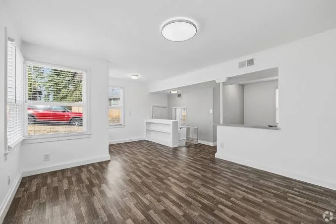 Empty living room with white walls, large windows, wood floor, and ceiling lights.