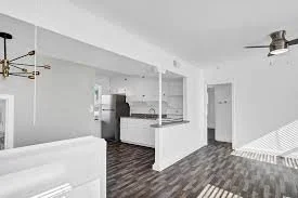 Empty living room with white walls, dark hardwood floors, and a ceiling fan, open to a kitchen area with white cabinets.