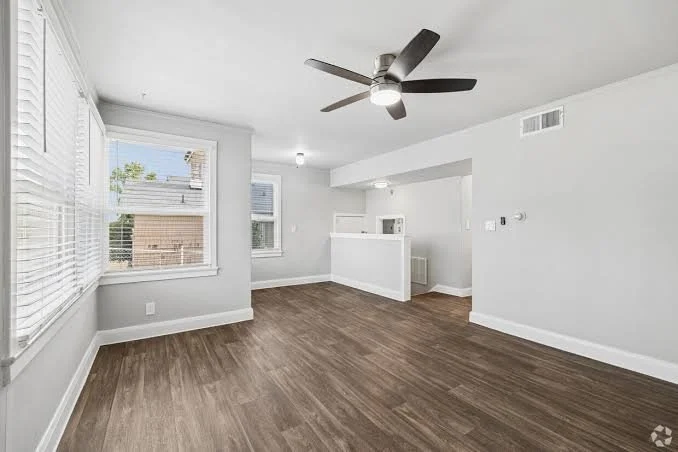 Empty living room with white walls, wooden flooring, large windows with blinds, ceiling fan, and a small partial wall with a microwave on it.