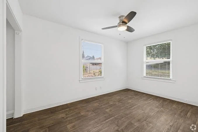 Empty room with white walls, two windows with blinds, hardwood floor, and a ceiling fan.