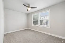 Empty room with white walls, a large window, ceiling fan, and light gray carpet.