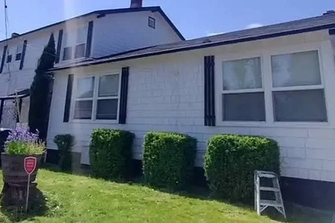 Front of a white house with four windows, surrounded by neatly trimmed bushes and a green lawn, with a partially visible front porch and a ladder near the house.