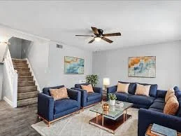Living room with two navy blue sofas, a glass coffee table, beige and pink pillows, a ceiling fan, and wall art with sea and landscape themes.