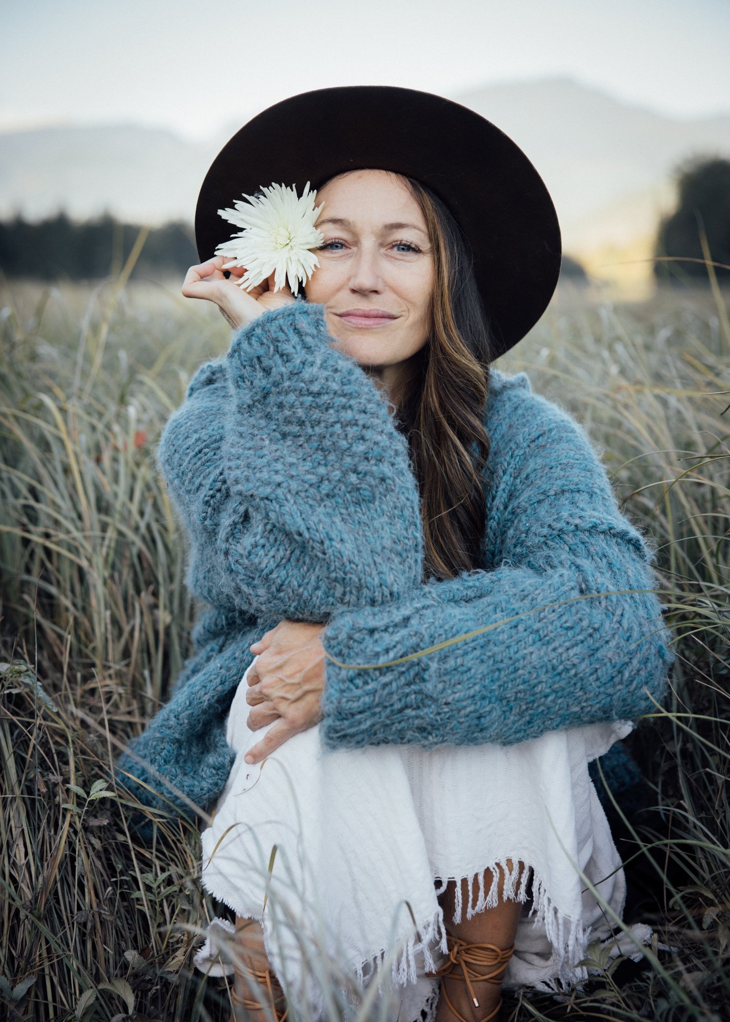 Woman wearing a black hat and blue sweater, seated in a grassy field and holding a white flower beside her face.