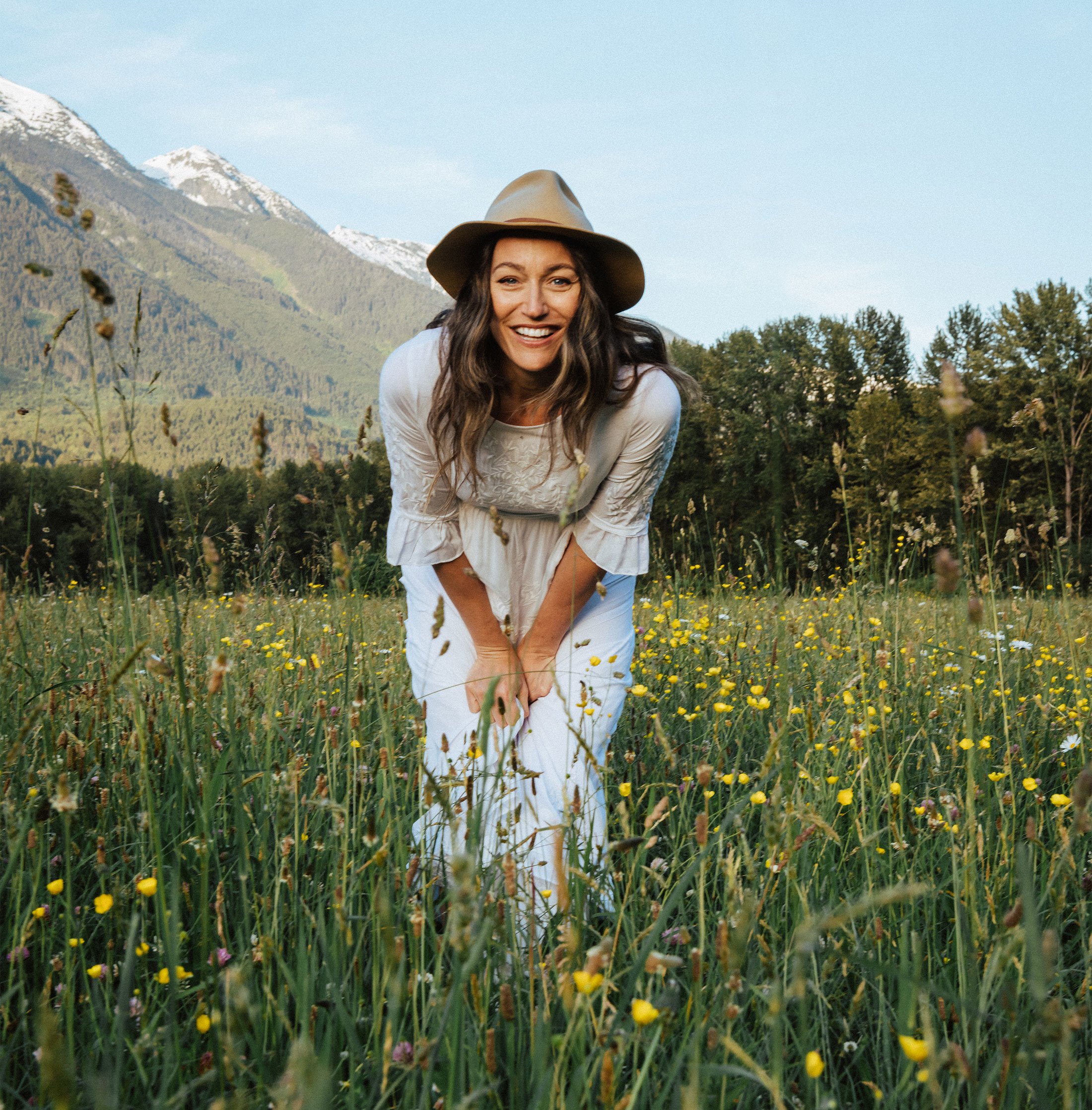 Woman in a white dress and wide-brimmed hat kneeling among wildflowers, with mountains in the background.
