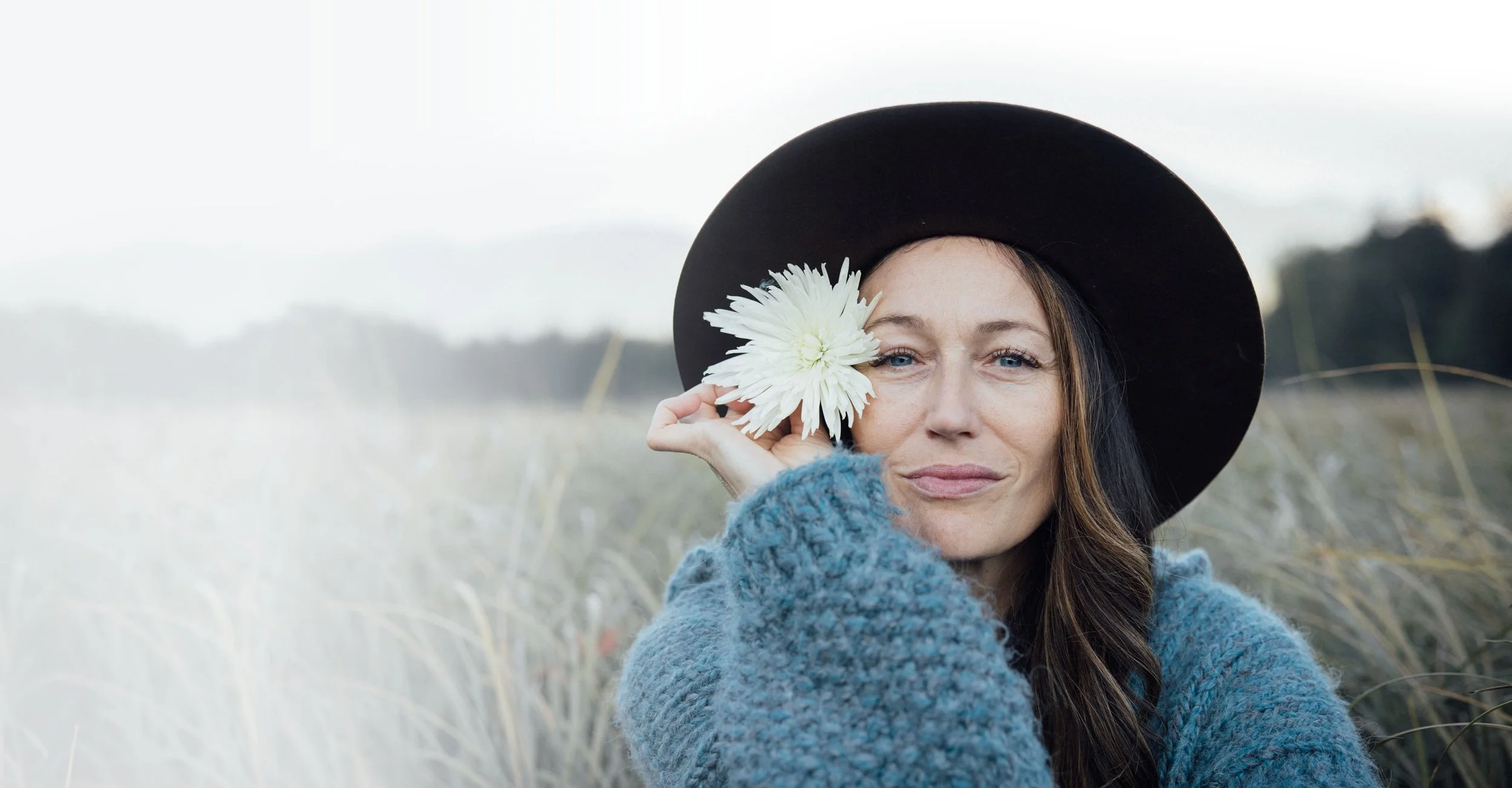Woman wearing a black hat and blue sweater, holding a white flower over one eye in a grassy field.