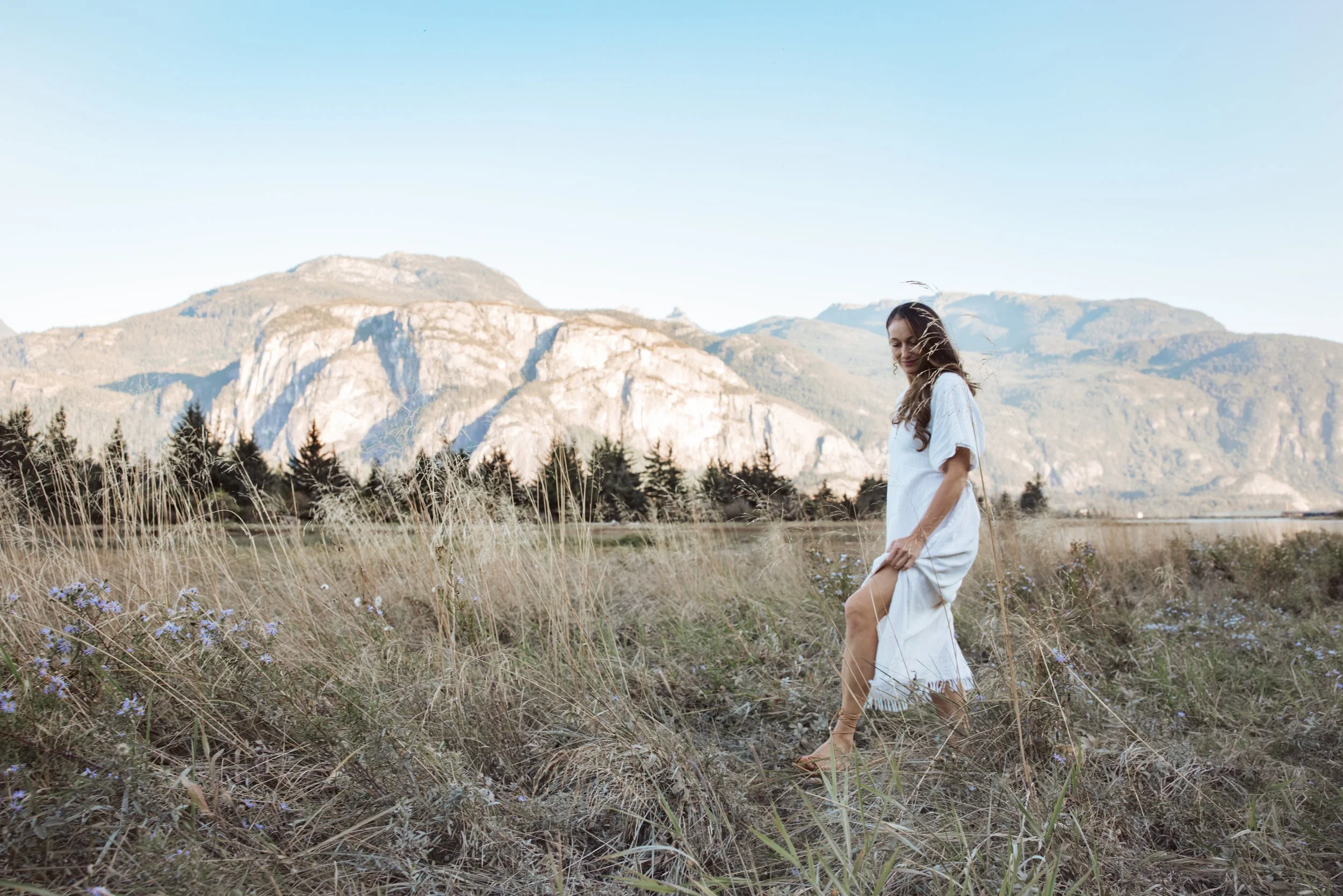 A young woman in a white dress walking through a field of tall dry grass and purple flowers, with mountains in the background under a clear blue sky.