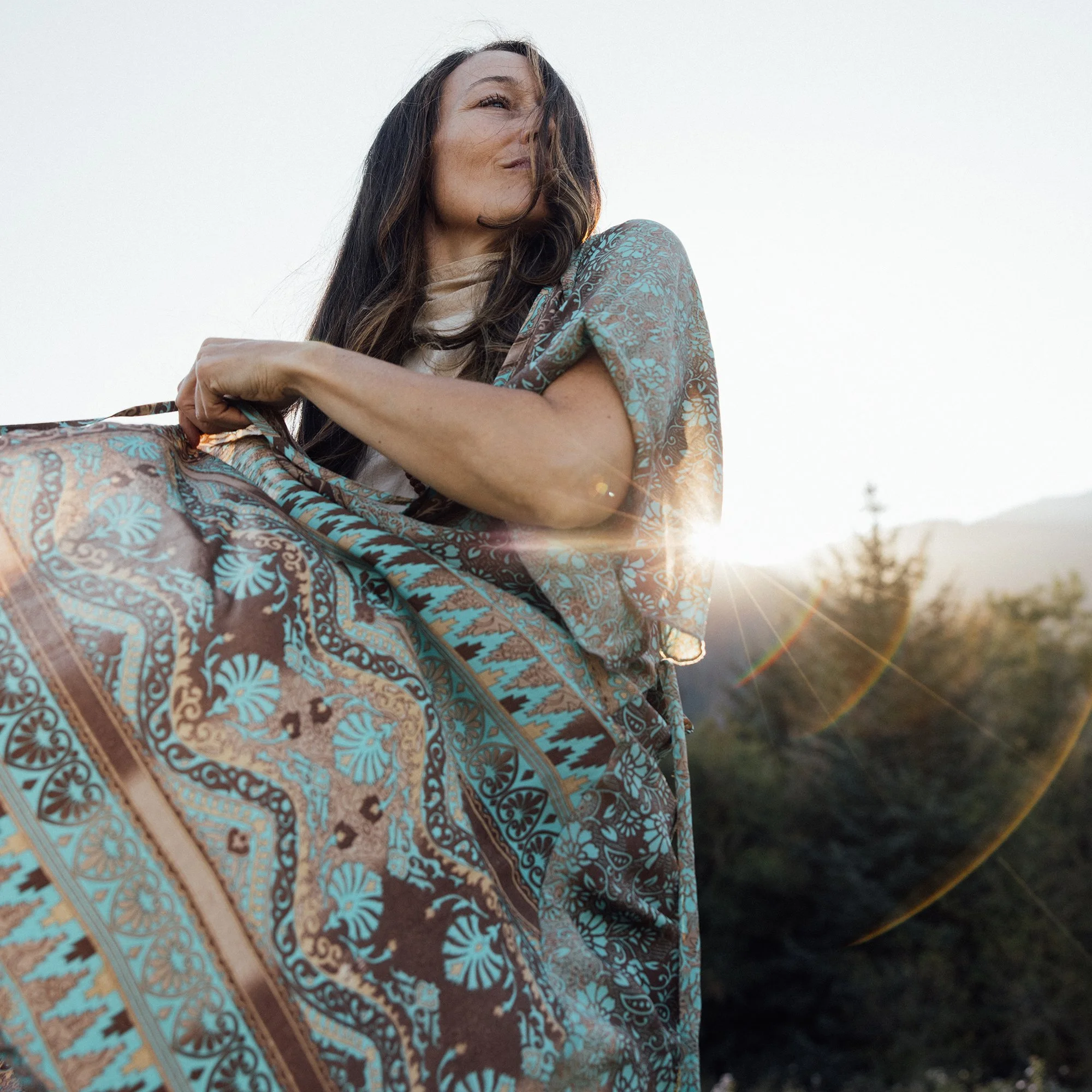 Woman with long dark hair wearing a flowing patterned dress, standing outdoors in warm sunlight.