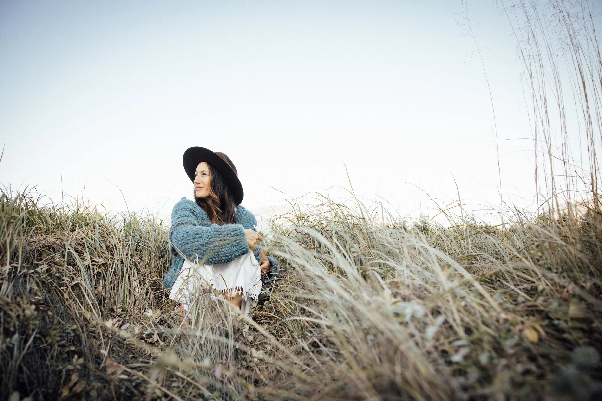Woman in a blue sweater and wide-brimmed hat sitting peacefully among tall grasses outdoors.