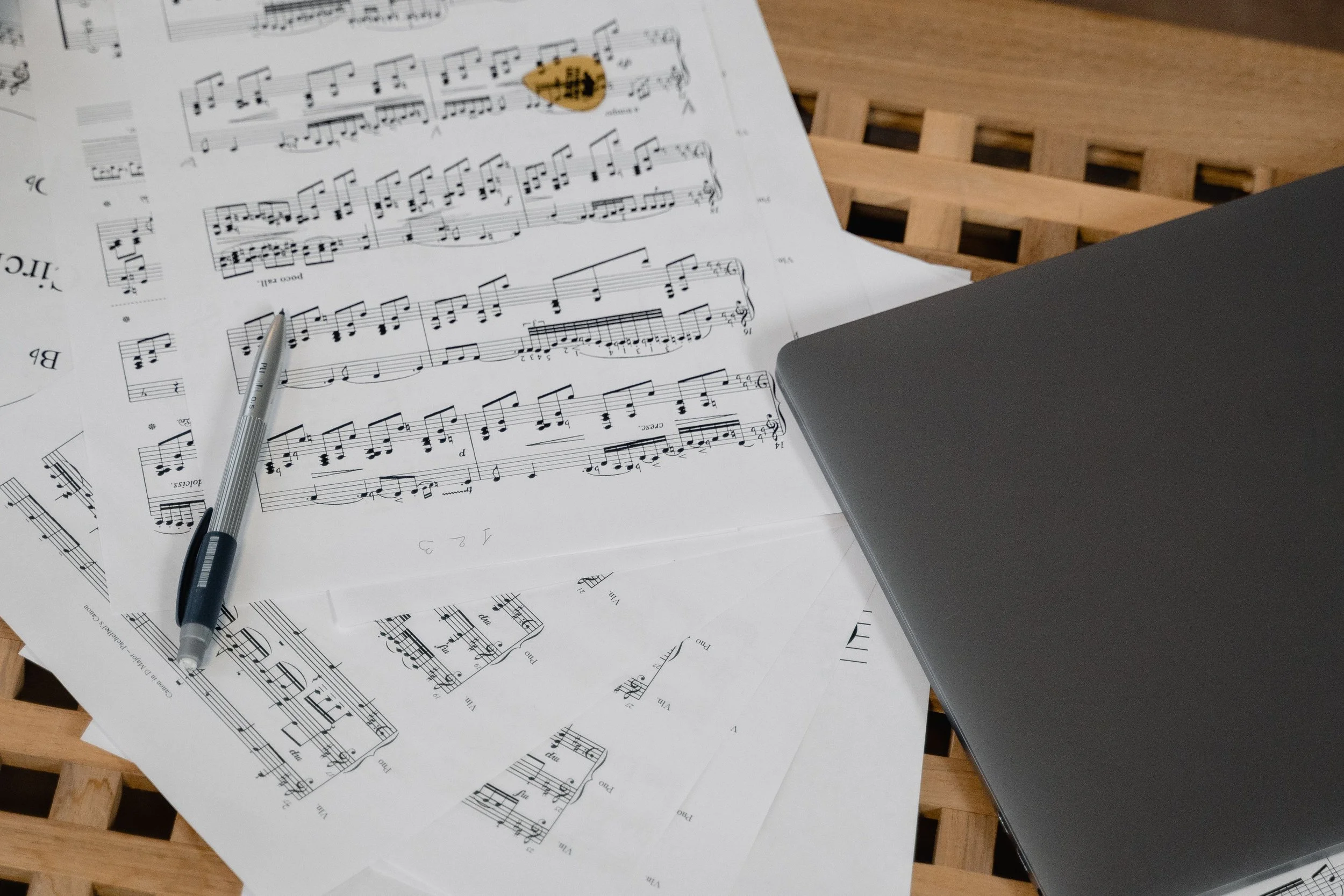 Sheets of printed sheet music, a pen, and a closed laptop on a wooden surface.