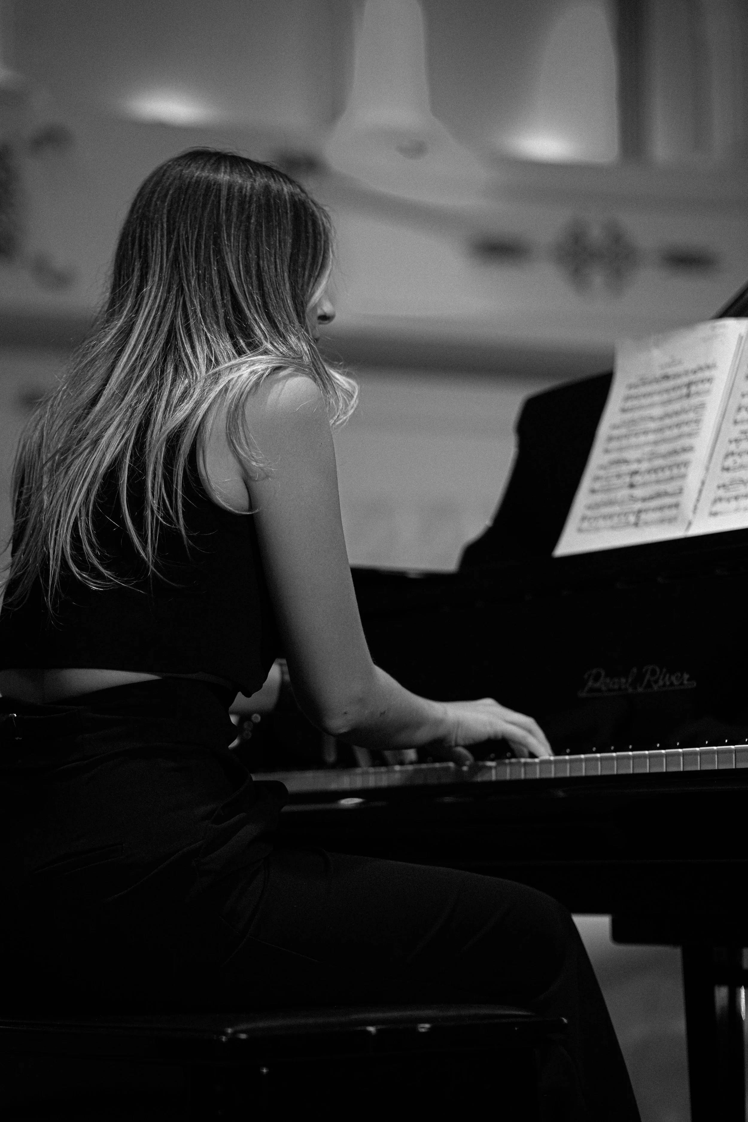 A woman with long wavy hair playing a grand piano, reading sheet music, in a room with a decorative ceiling.