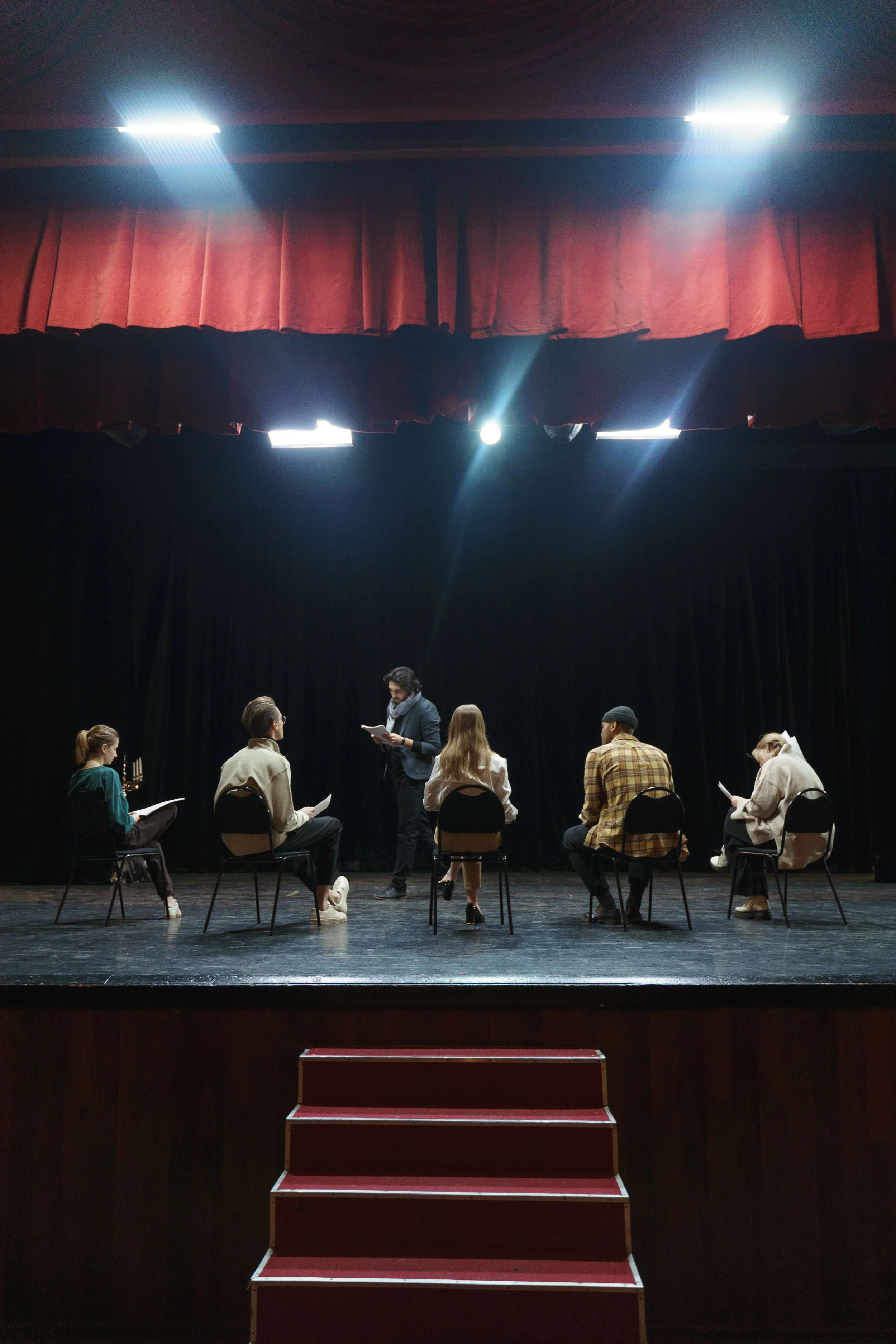 People on stage rehearsing, seated in a circle, with a person standing and reading from paper, in front of a red curtain and stage lights