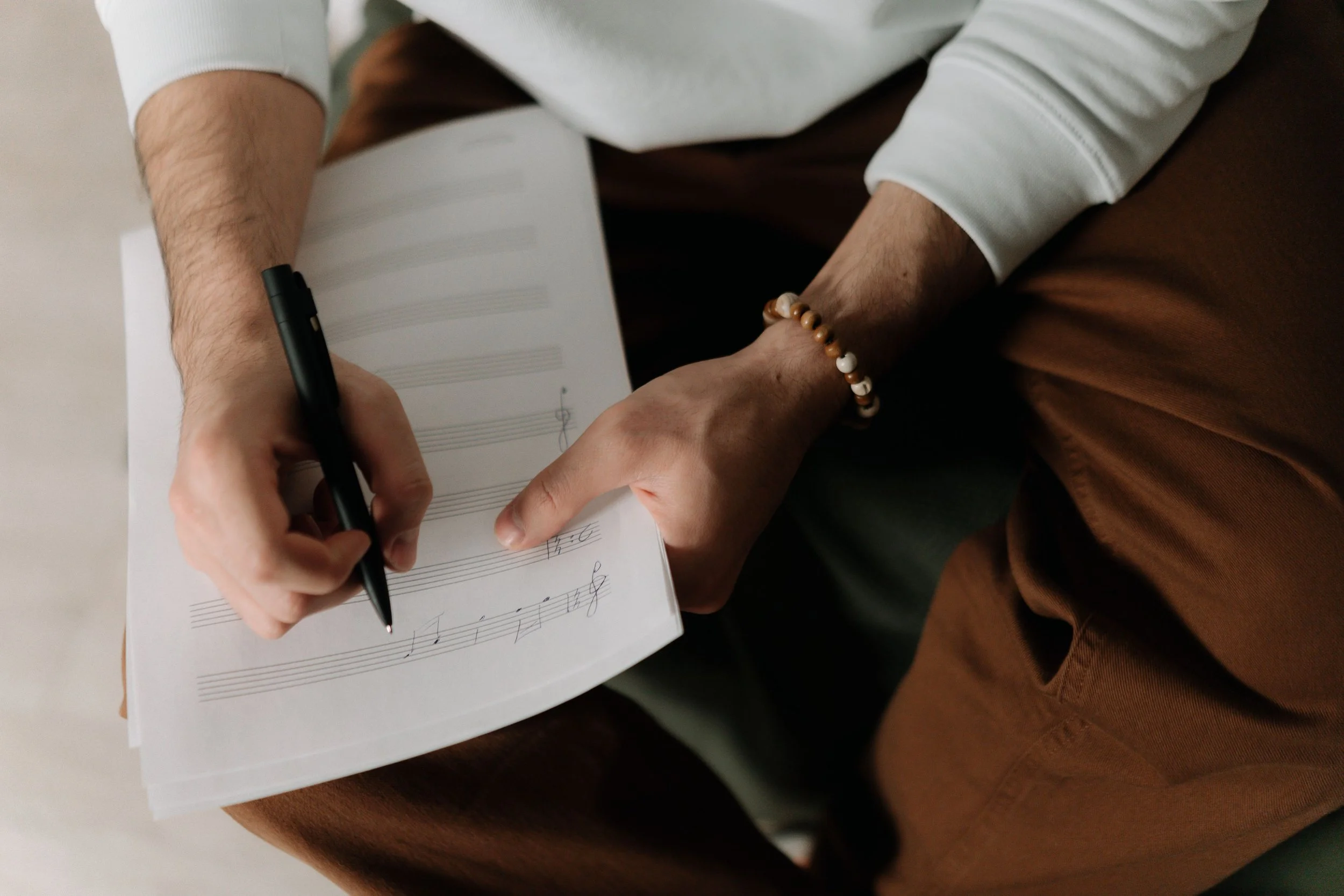 A person wearing a white sleeve writing music notation on sheet music with a black pen, while their other hand holds the sheet, sitting on a wooden surface.