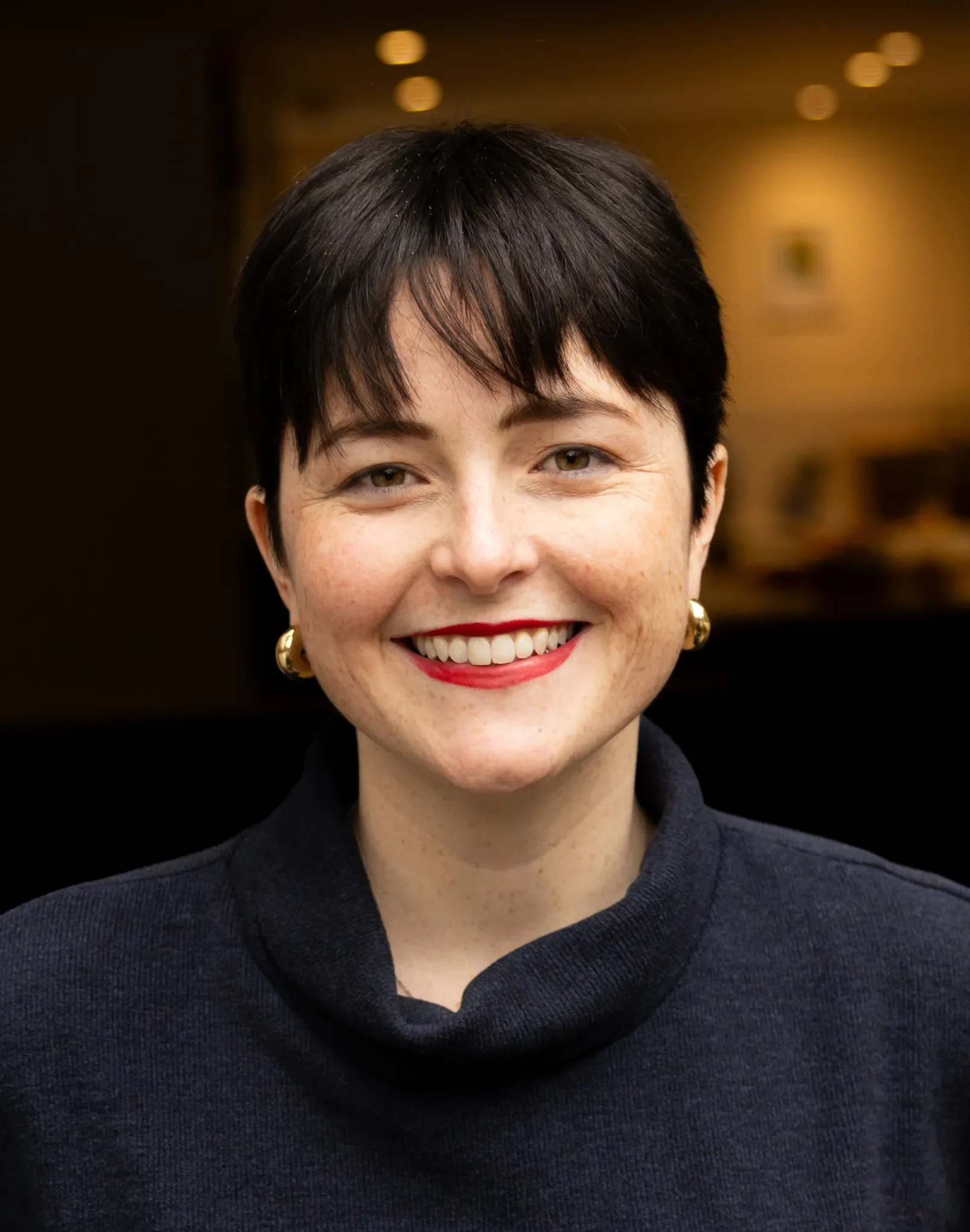 Close-up of a woman with short dark hair and green eyes, smiling, wearing a dark sweater and gold earrings, with a blurred background featuring warm lighting.