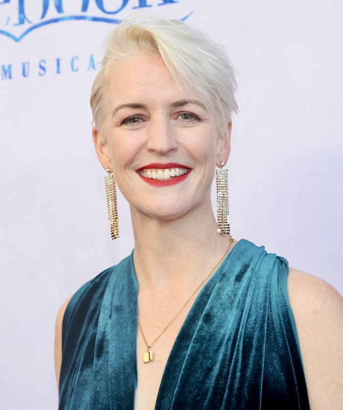 A woman with short, platinum blonde hair and red lipstick smiling at the camera, wearing long chandelier earrings, a gold necklace with a small pendant, and a teal velvet dress.