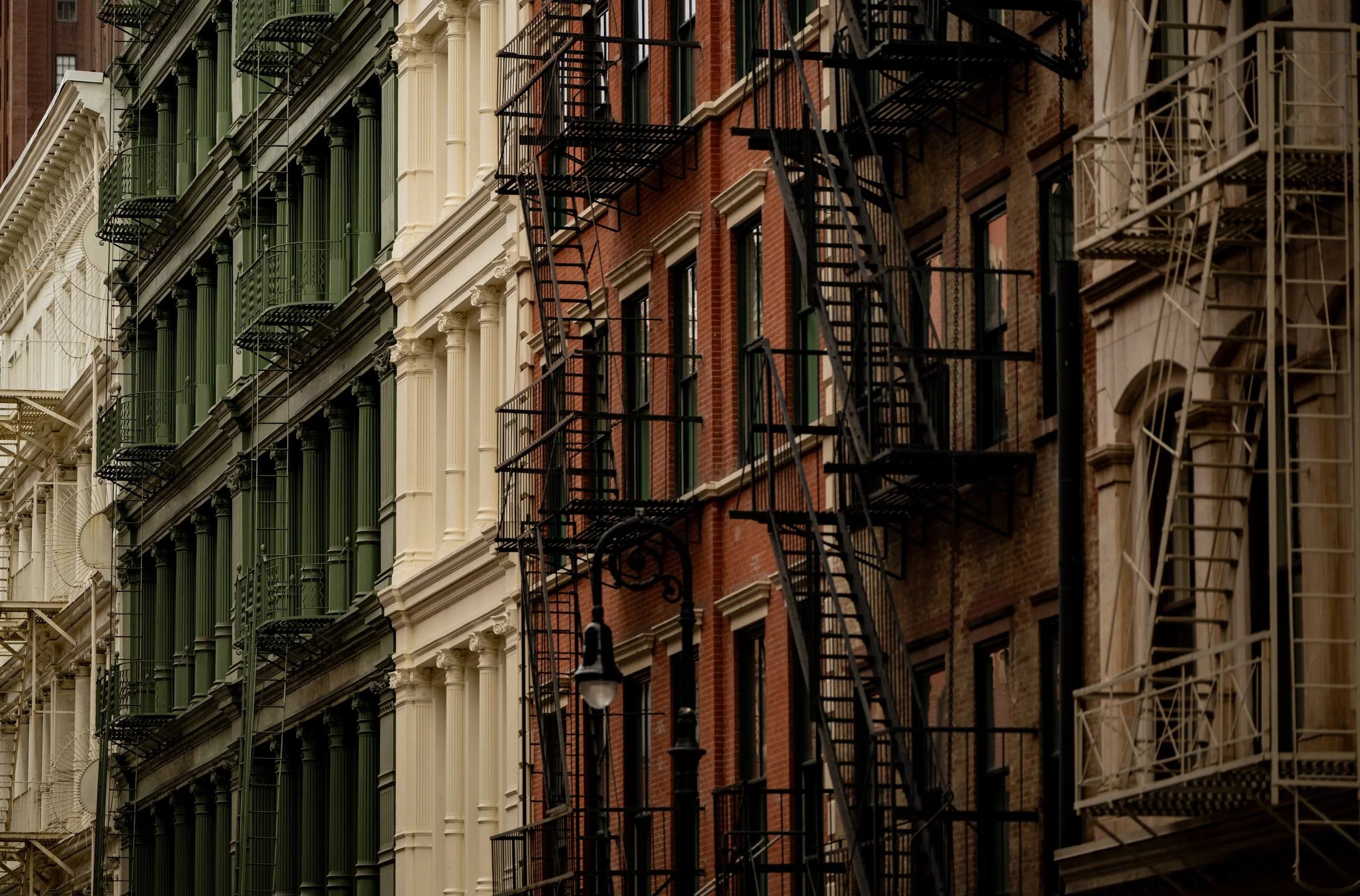 Multiple adjacent city buildings with fire escapes on their facades, painted in green, white, red brick, and beige, in a city street.