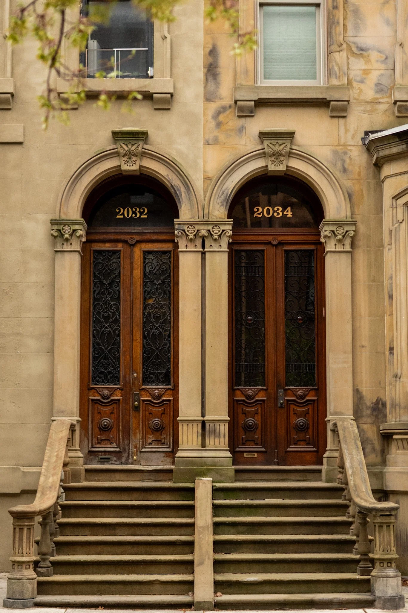 Double wooden doors with decorative ironwork, arched windows above labeled 2032 and 2034, stone staircase and railing, stone building facade with window on second floor.