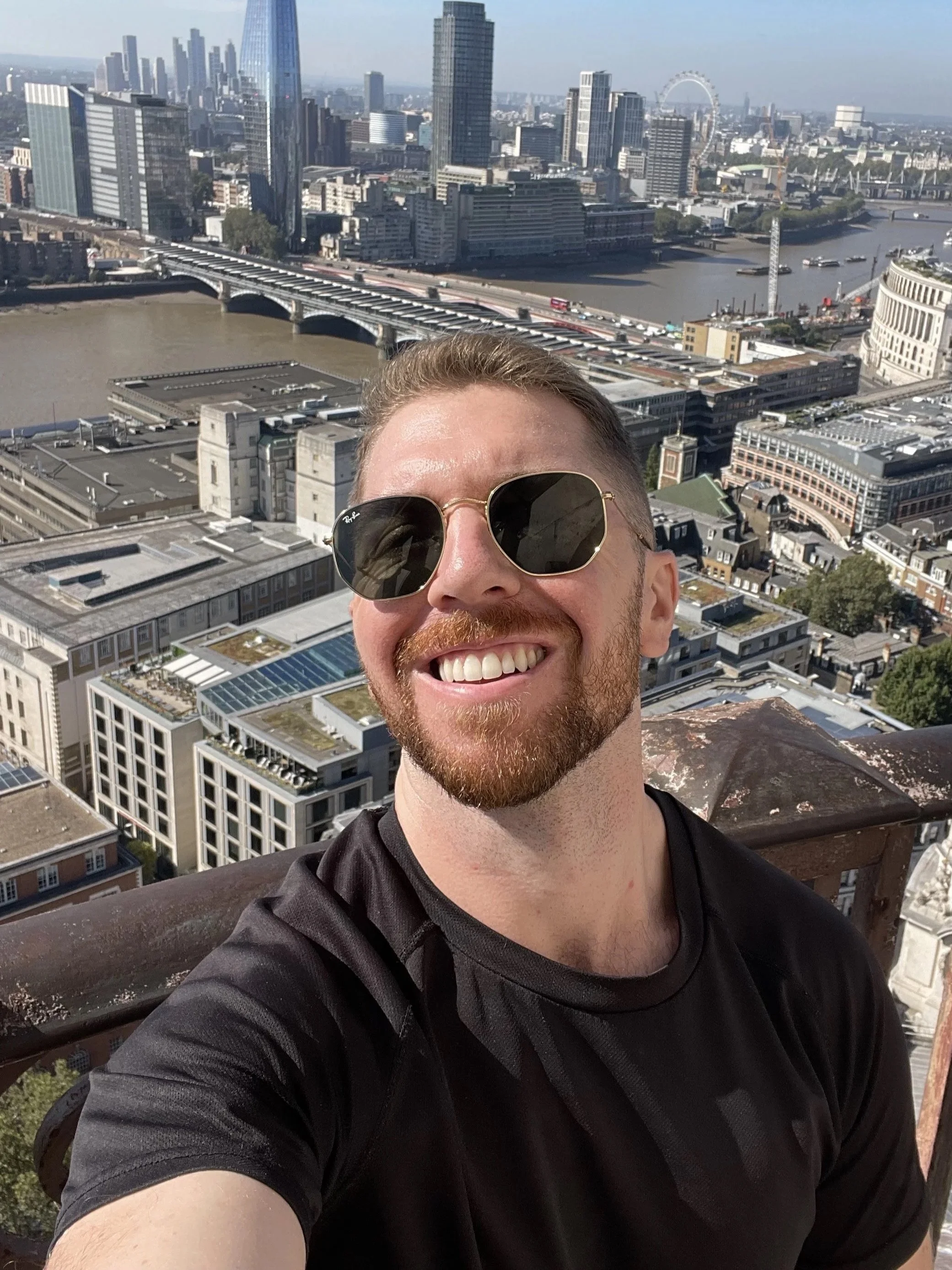 A man with sunglasses smiling for a selfie on a high rooftop, with the London skyline and the River Thames visible in the background.