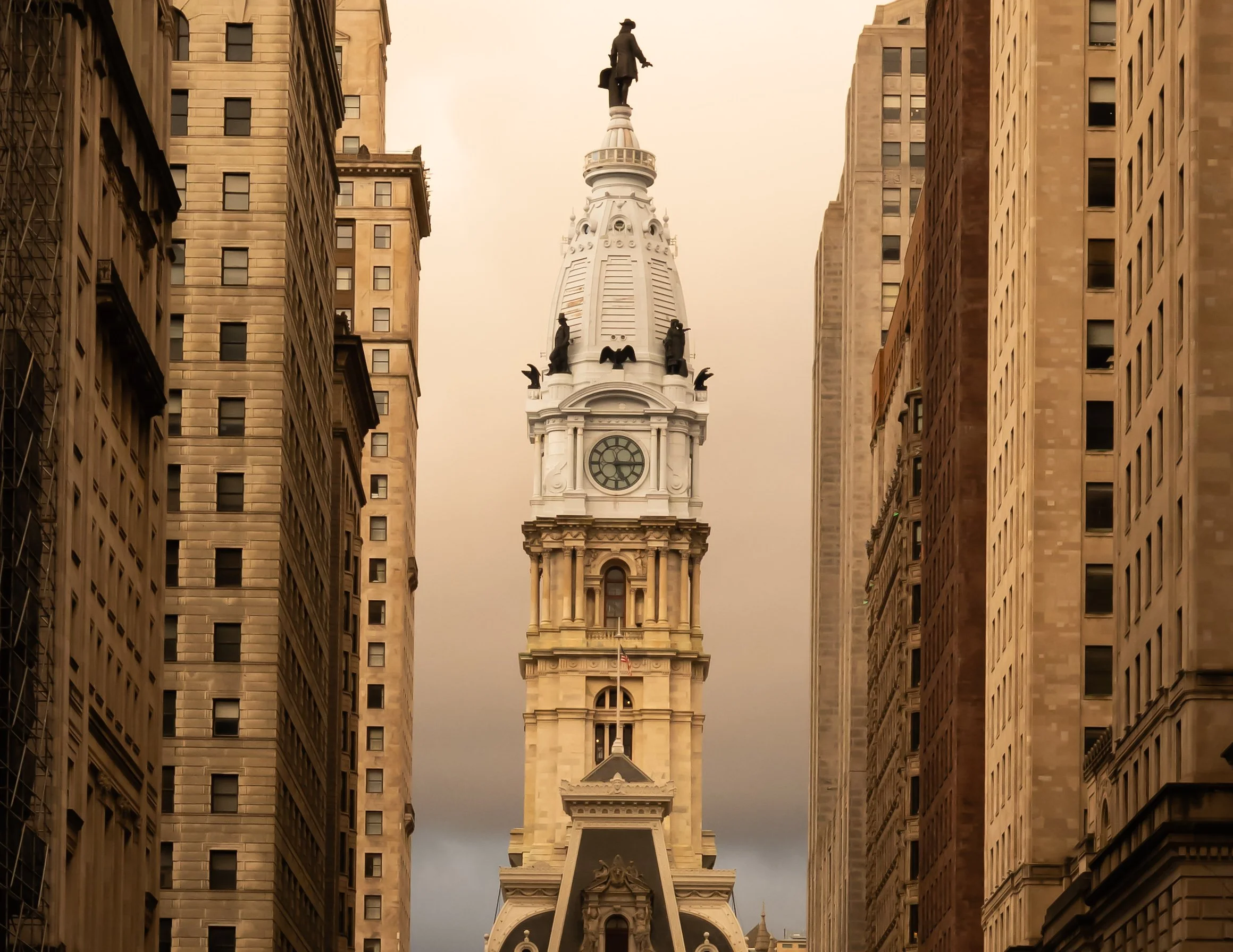 A clock tower with a statue of a man in a coat and hat on top, between two tall, beige city buildings.