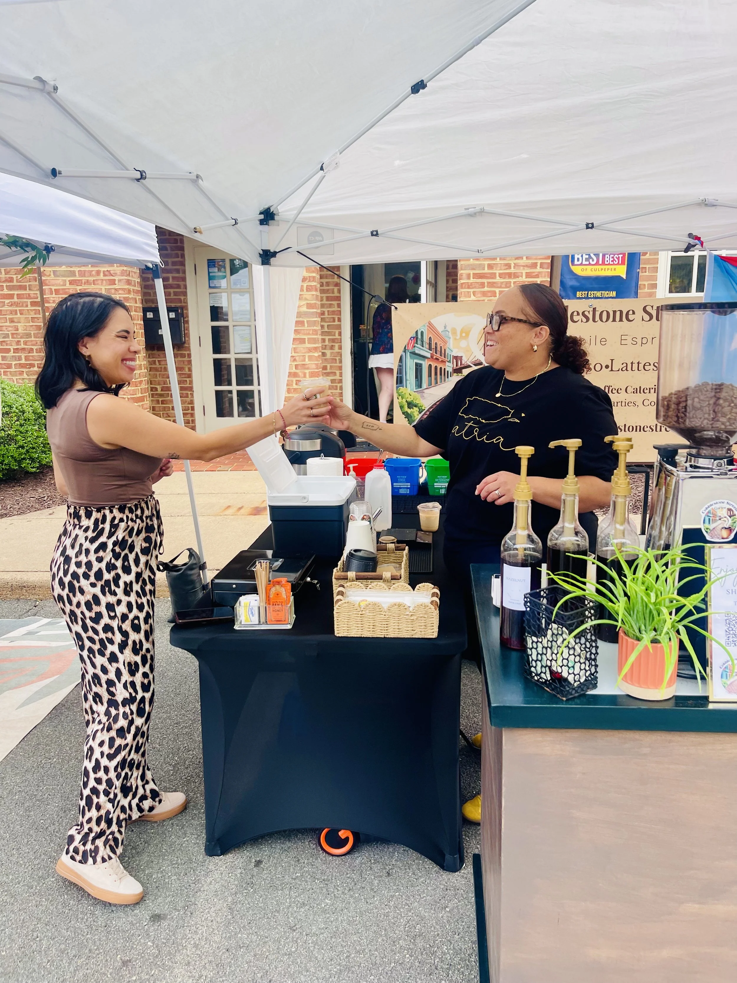 Two women exchanging a handshake at an outdoor market stand. One woman is wearing animal print pants and a sleeveless top, smiling, while the other woman is wearing glasses and a black shirt with gold lettering, also smiling. The stand has various items including a basket, bottles, and a coffee machine, with a banner and a plant on the right side.